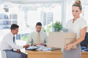 sad woman carrying box in an office with two men sitting at a desk behind her