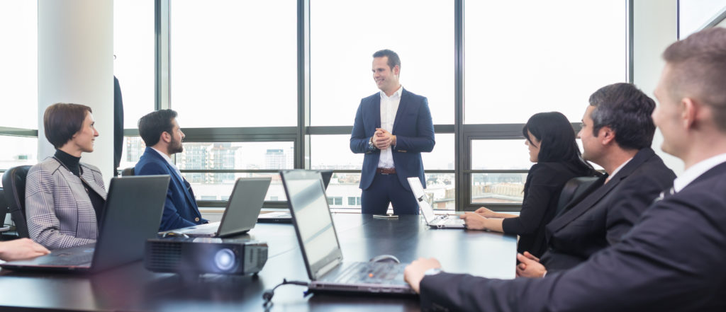 an office with employees on laptops and their manager standing up talking to them